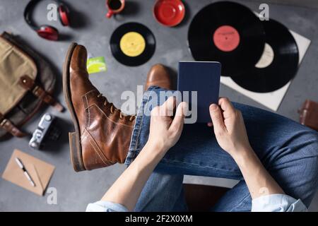 Herren Hand hält Pass oder Notebook und Bein in Jeans mit alten Reisen Lederstiefel Schuhe. Entspannung und Kunst Musik Lifestyle Konzept Stockfoto