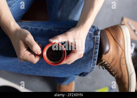 Herren Hand hält Tasse Kaffee und Bein in Jeans mit alten Reisen Lederstiefel Schuhe. Entspannung und Kunst Musik Lifestyle Konzept Stockfoto