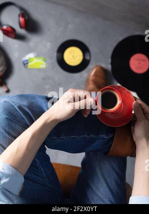 Herren Hand hält Tasse Kaffee und Bein in Jeans mit alten Reisen Lederstiefel Schuhe. Entspannung und Kunst Musik Lifestyle Konzept Stockfoto