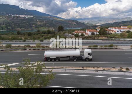 Tankwagen auf der Autobahn mit einer Landschaft aus Bergen, Wolken, einem Stausee und einer Stadt im Hintergrund. Stockfoto