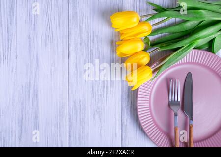 Draufsicht auf eine Komposition aus einem Bouquet aus gelben Tulpen, einem rosa Teller mit Besteck auf hellem Holzhintergrund. Selektiver Fokus. Speicherplatz für Ihre te kopieren Stockfoto