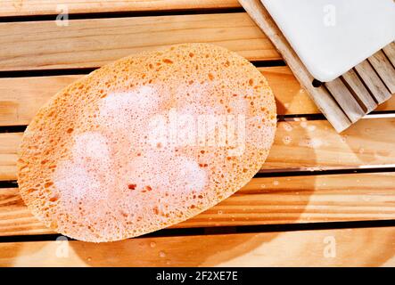 Orangefarbener Badeschwamm mit Blasen auf Holzgrund, im Hintergrund weiße Seife Stockfoto
