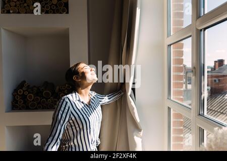 Junge kaukasische Frau öffnet die Vorhänge und schaut auf die Fenster an einem sonnigen Morgen Stockfoto