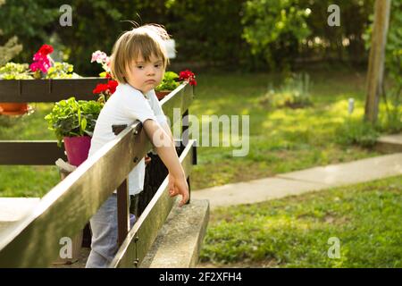 Portrait von niedlichen kleinen Jungen mit Down-Syndrom lehnte sich auf Zaun beim Genießen und Entspannen Terrasse während Quarantäne. Stockfoto