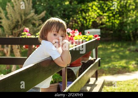 Portrait von niedlichen kleinen Jungen mit Down-Syndrom lehnte sich auf Zaun beim Genießen und Entspannen Terrasse während Quarantäne. Stockfoto