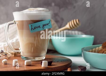 Kaffee Latte Tasse mit Love You Aufkleber auf Holztablett Mit türkisfarbenen Schalen mit Plätzchen und Marshmallows an der grauen Wand Hintergrund Stockfoto