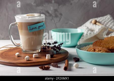 Kaffee Latte Tasse mit Love You Aufkleber auf Holztablett Mit türkisfarbenen Schalen mit Plätzchen und Marshmallows an der grauen Wand Hintergrund Stockfoto