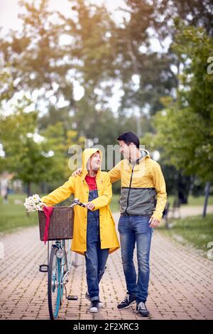 Vertikales Bild von schönen weiblichen Blick mit Liebe und Zuneigung kaukasischer junger Mann umarmt sie beim Anschieben des Fahrrads Ein regnerischer Tag im Park Stockfoto