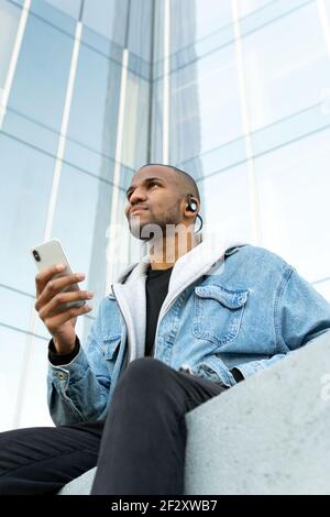 Von unten der erwachsenen ethnischen männlichen Hören zu Lied aus Kopfhörer während man mit Handy in der Stadt sitzt und sich freut Stockfoto