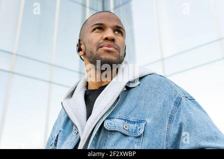 Von unten von erwachsenen bärtigen ethnischen Männern in drahtlosen Ohrhörern Beim Blick in die Stadt dem Lied lauschen Stockfoto