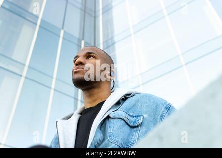 Von unten von erwachsenen bärtigen ethnischen Männern in drahtlosen Ohrhörern Beim Blick in die Stadt dem Lied lauschen Stockfoto