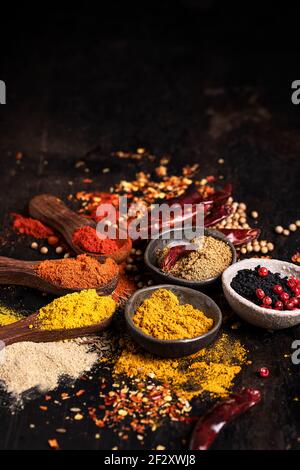 Verschiedene aromatische bunte Gewürze in Schalen und Holzlöffel angeordnet Auf dunklem Tisch mit verstreuten Gewürzen Stockfoto