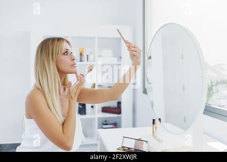 Seitenansicht des Weibchens mit perfekter Haut Auftragen Puder und Berühren des Gesichts während der Selbstaufnahme auf dem Smartphone während der Arbeit Make-up zu Hause Stockfoto