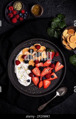 Blick von oben auf leckere gesunde hausgemachte Frühstück mit frischen Auswahl Beeren serviert mit Joghurt und Cookies in schwarzer Platte auf Tisch mit Zutaten Stockfoto