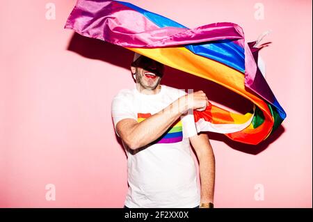 Fröhlich unrasiert homosexuellen Mann mit roten Lippen in weiß t shirt lächelnd, während die LGBT-Flagge vor rosa Hintergrund winkt Stockfoto
