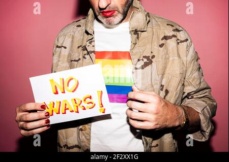 Crop anonymen bärtigen exzentrischen Mann mittleren Alters im Hemd mit LGBT-Flagge mit Make-up und Maniküre zeigt Papier mit Nein Kriege Inschrift gegen Rosa Stockfoto