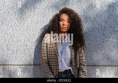 Nachdenkliche afroamerikanische Frau im trendigen Outfit mit lockigen Haaren Blick auf die Kamera, während Sie auf der Straße in der Nähe einer Betonwand stehen Stockfoto