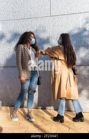 Seitenansicht der ethnischen weiblichen Freunde in legerer Kleidung mit Medizinische Masken, die einander anblicken, während sie mit Ellbogen grüßen Stockfoto
