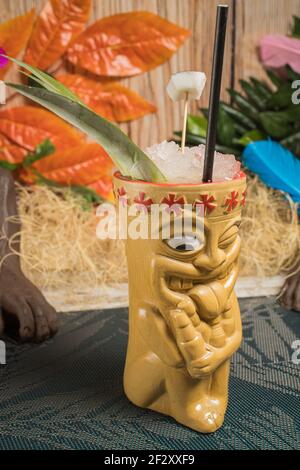 Polynesische Tiki-Tasse mit kaltem Alkoholgetränk, dekoriert mit Stroh Und grüne Blätter gegen bunte Blätter und trockenes Gras gelegt Stockfoto