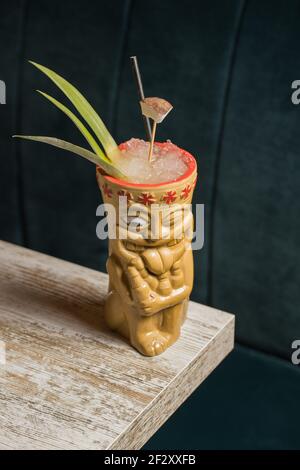 Polynesische Tiki-Tasse mit kaltem Alkoholgetränk, dekoriert mit Stroh Und grüne Ananasblätter gegen auf Holztisch gelegt Stockfoto