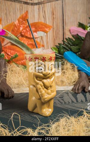 Polynesische Tiki-Tasse mit kaltem Alkoholgetränk, dekoriert mit Stroh Und grüne Blätter gegen bunte Blätter und trockenes Gras gelegt Stockfoto