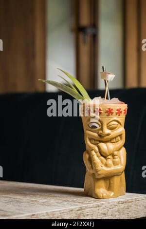 Polynesische Tiki-Tasse mit kaltem Alkoholgetränk, dekoriert mit Stroh Und grüne Ananasblätter gegen auf Holztisch gelegt Stockfoto