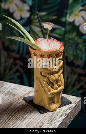 Polynesische Tiki-Tasse mit kaltem Alkoholgetränk, dekoriert mit Stroh Und grüne Ananasblätter gegen auf Holztisch gelegt Stockfoto