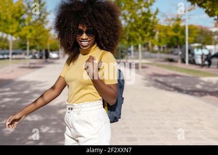 Fröhliche junge afroamerikanische Reisende mit Rucksack stehen auf Gehweg in der Stadt an sonnigen Tag Stockfoto