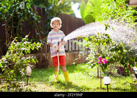 Kinder bewässern Blumen und Pflanzen im Garten. Kind mit Wasserschlauch in sonnigen blühenden Hinterhof. Kleiner Junge im Garten. Sommerspaß im Freien zu Hause. Stockfoto