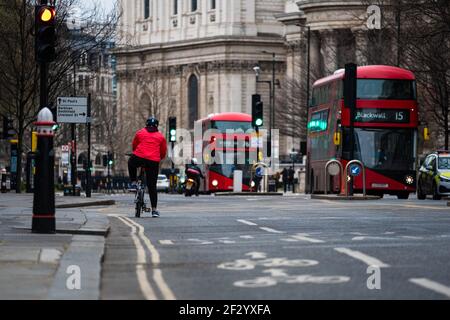 Radfahrer mit roter Jacke warten auf die Ampel an einer Kreuzung, zwei Doppeldeckerbusse fahren im Hintergrund vorbei. Straßen von londo Stockfoto