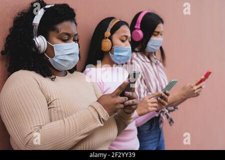 Frauen mit mehreren Rassen, die ein Smartphone nutzen und kabellos Musik hören Kopfhörer, während die Gesichtsmaske während der Corona-Virus-Pandemie getragen wird Stockfoto