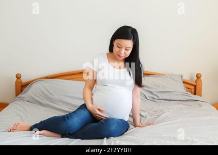 Lächelnde asiatische chinesische schwangere Frau auf einem Bett sitzend berühren Bauch. Junge Dame in weißen T-Shorts und blauen Jeans zu Hause erwartet. Gesund glücklich Stockfoto