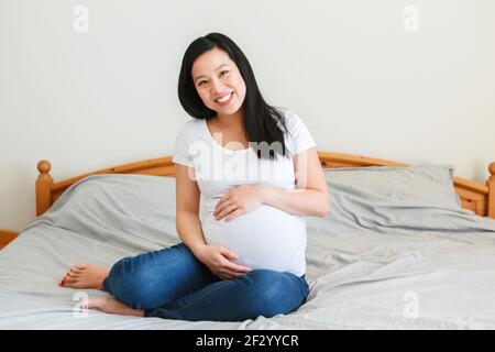 Lächelnde asiatische chinesische schwangere Frau auf einem Bett sitzend berühren Bauch. Junge Dame in weißen T-Shorts und blauen Jeans zu Hause erwartet. Gesund glücklich Stockfoto