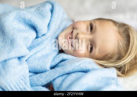 Niedliche kleine kaukasische Mädchen sieben Jahre alt mit blonden Haaren liegen auf dem Bett zu Hause im Schlafzimmer. Kind trägt modisch gemütlich Strickpullover blau Farbe Stockfoto