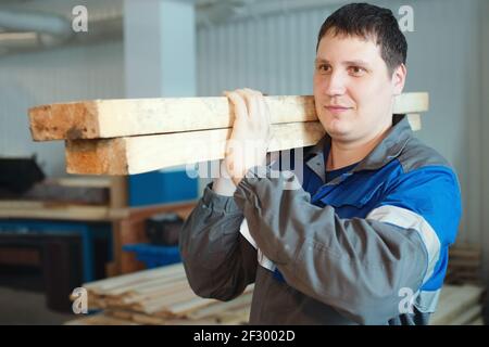 Ein Sägearbeiter in Overalls trägt Holzstangen auf seiner Schulter. Produktion und Lieferung von Baustoffen. Stockfoto