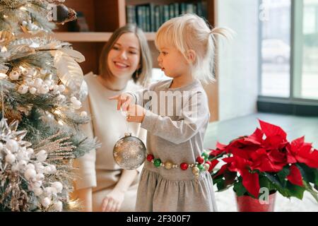 Glückliche kaukasische Mutter mit Baby Mädchen Dekoration Weihnachtsbaum mit Ornamenten Kugeln Spielzeug. Glückliche Familie feiert Weihnachten oder Neujahr Winterurlaub Stockfoto