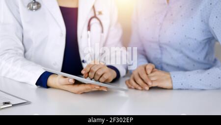 Unbekannte Ärztin zeigt ihrem Patienten eine Beschreibung der Medikamente, während sie zusammen am Schreibtisch in der sonnigen Vitrine in einer Klinik sitzt Stockfoto