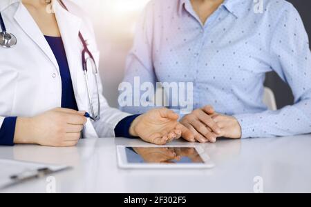 Unbekannte Ärztin zeigt ihrem Patienten eine Beschreibung der Medikamente, während sie zusammen am Schreibtisch in der sonnigen Vitrine in einer Klinik sitzt Stockfoto