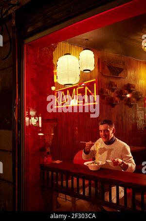 Ein europäischer Mann, der Ramen vor der Ladenfront eines japanischen Restaurants am Abend isst. Stockfoto