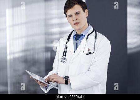 Ein junger, freundlicher Arzt überprüft einige Informationen auf seinem Computer-Tablet. Porträt eines Facharztes bei der Arbeit in einer Klinik. Medizinkonzept Stockfoto