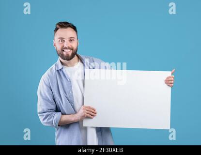 Fröhlich kaukasischen Mann demonstriert weiße leere Plakat auf blauem Studio-Hintergrund, mockup für Design Stockfoto