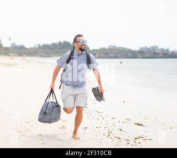 Fröhlicher erwachsener Mann mit Tasche, der sich am tropischen Strand amüsieren kann Stockfoto