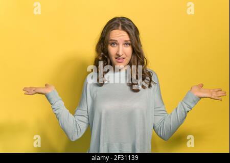 Unsicherheitskonzept. Eine junge lockige Brünette Frau spreizt ihre Arme und zuckt Schultern, weiß nicht, was zu tun ist, eine Frau in Zweifel verloren, Studio erschossen auf gelb Stockfoto