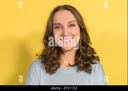 Nahaufnahme Porträt der attraktiven jungen Brünette Frau, eine blauäugige Frau mit natürlicher Schönheit schaut auf die Kamera und lacht, isoliert auf gelbem Hintergrund Stockfoto