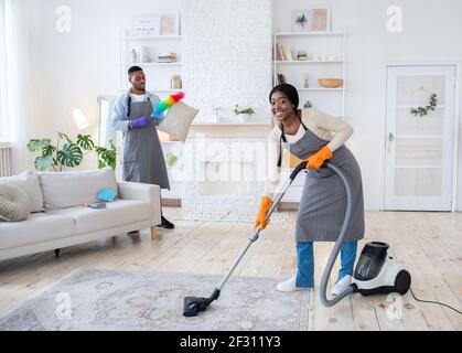 Glückliche schwarze Frau Staubsaugen Boden, während ihr Freund Staubkissen in ihrer Wohnung, kopieren Raum Stockfoto