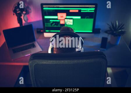 Kaukasische Männer in seinem 40s trägt Kopfhörer vor seinem Desktop-Büro-Computer. Rückansicht Des Stuhls. Moderne Technologien und Home Office-Arbeit. Stockfoto