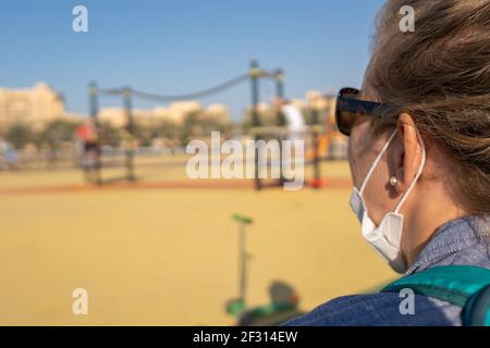 Über der Schulter einer Mutter mit COVID-19 Maske und Blick auf Kinder auf dem Spielplatz in der Sonne, kopieren Raum und verschwommener Hintergrund. Stockfoto