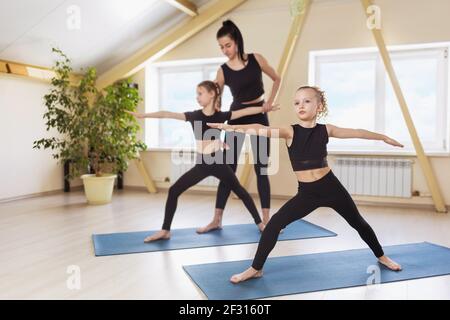 Kleine Mädchen in schwarzer Sportkleidung Yoga üben führen Virabhadrasana, die Krieger Pose, unter der Anleitung ihres Trainers. Gruppenschulungskonzept Stockfoto