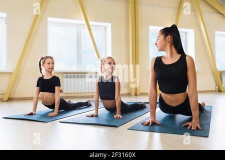 Eine Trainerin und zwei kleine Mädchen, die im Studio Yoga praktizieren, machen eine Übung mit einem Hund, der sich auf Gymnastikmatten zeigt, Urdhva Mukha Shvanasana p Stockfoto