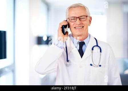 Aufnahme von glücklichen männlichen Arzt mit seinem Handy und im Gespräch mit jemandem, während im Zimmer des Docto stehen. Stockfoto